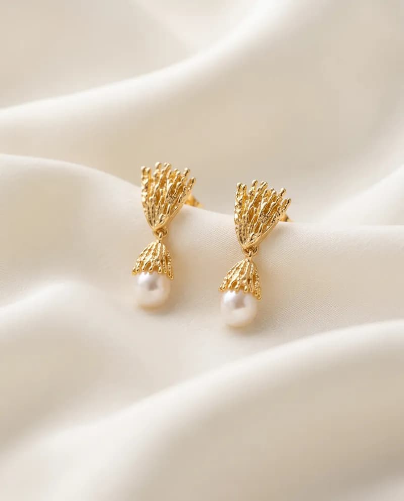 Pendientes de ramas de coral en baño de oro abiertas hacia arriba, con una pequeña perla de cristal colgando, sobre satén marfil