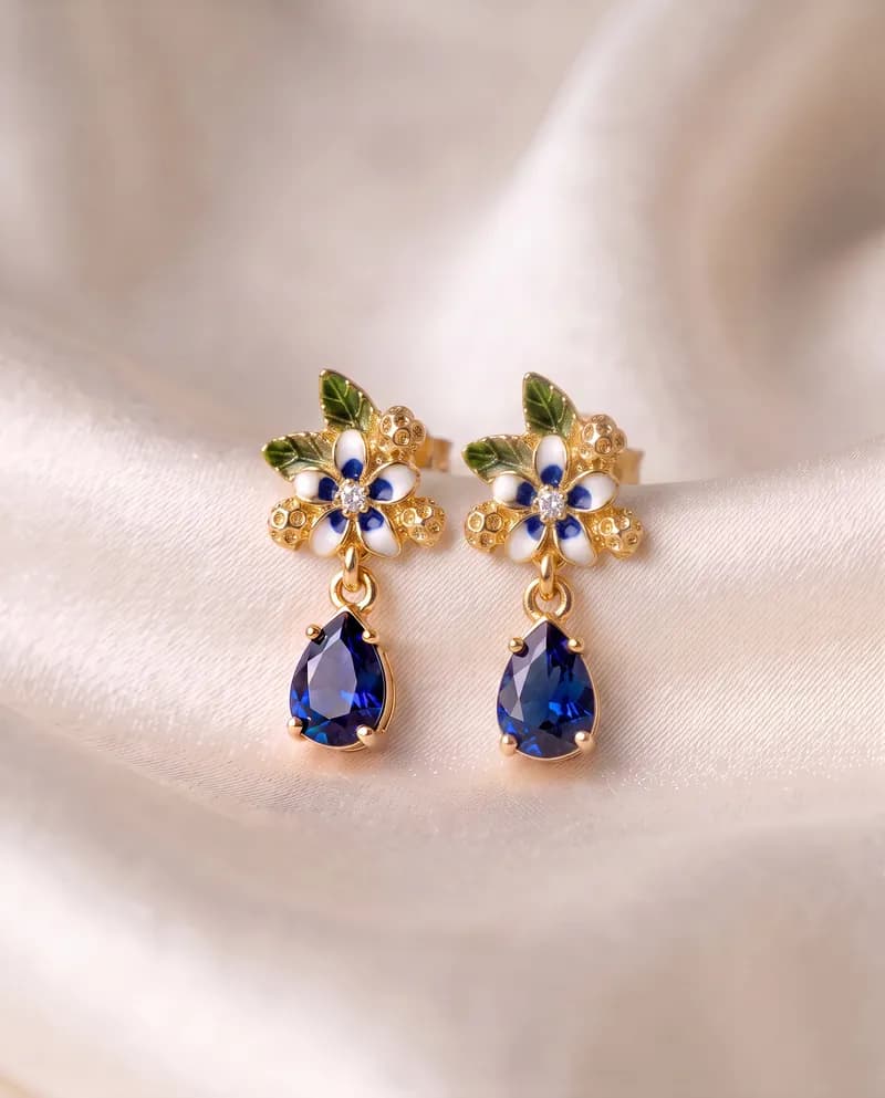Pendientes con una flor esmaltada a mano en blanco y azul, hojas verdes y una gota azul facetada colgando debajo, en baño de oro